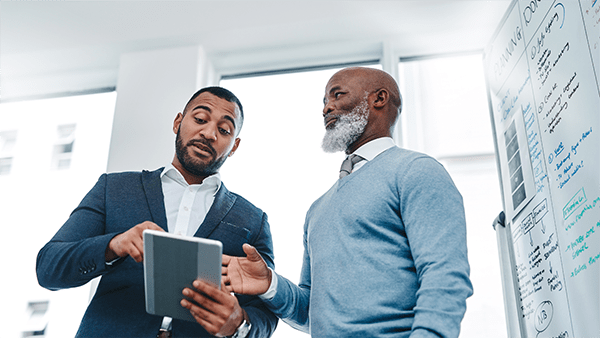 Two men in a modern office discuss a project. One holds a tablet, and the other gestures toward a whiteboard filled with notes and diagrams, conveying collaboration.