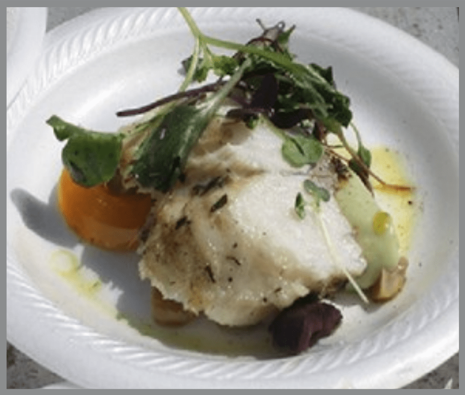 Un plato servido que presenta un filete de pescado blanco cubierto con hierbas y hojas verdes, servido en un plato blanco acanalado con una salsa ligera. El pescado descansa sobre una base de verduras, incluyendo un trozo visible de calabaza de color naranja, con pequeños adornos y hojas verdes colocados encima.