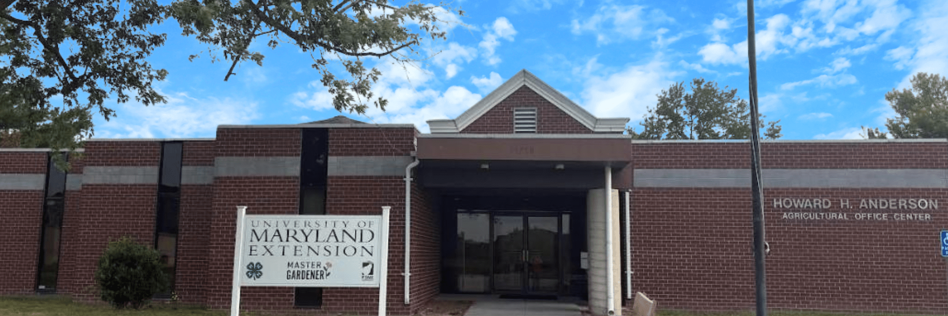 Brick building with white University of Maryland Extension sign to left of door entrance, flag pole to the right, blue sky with white clouds in background.