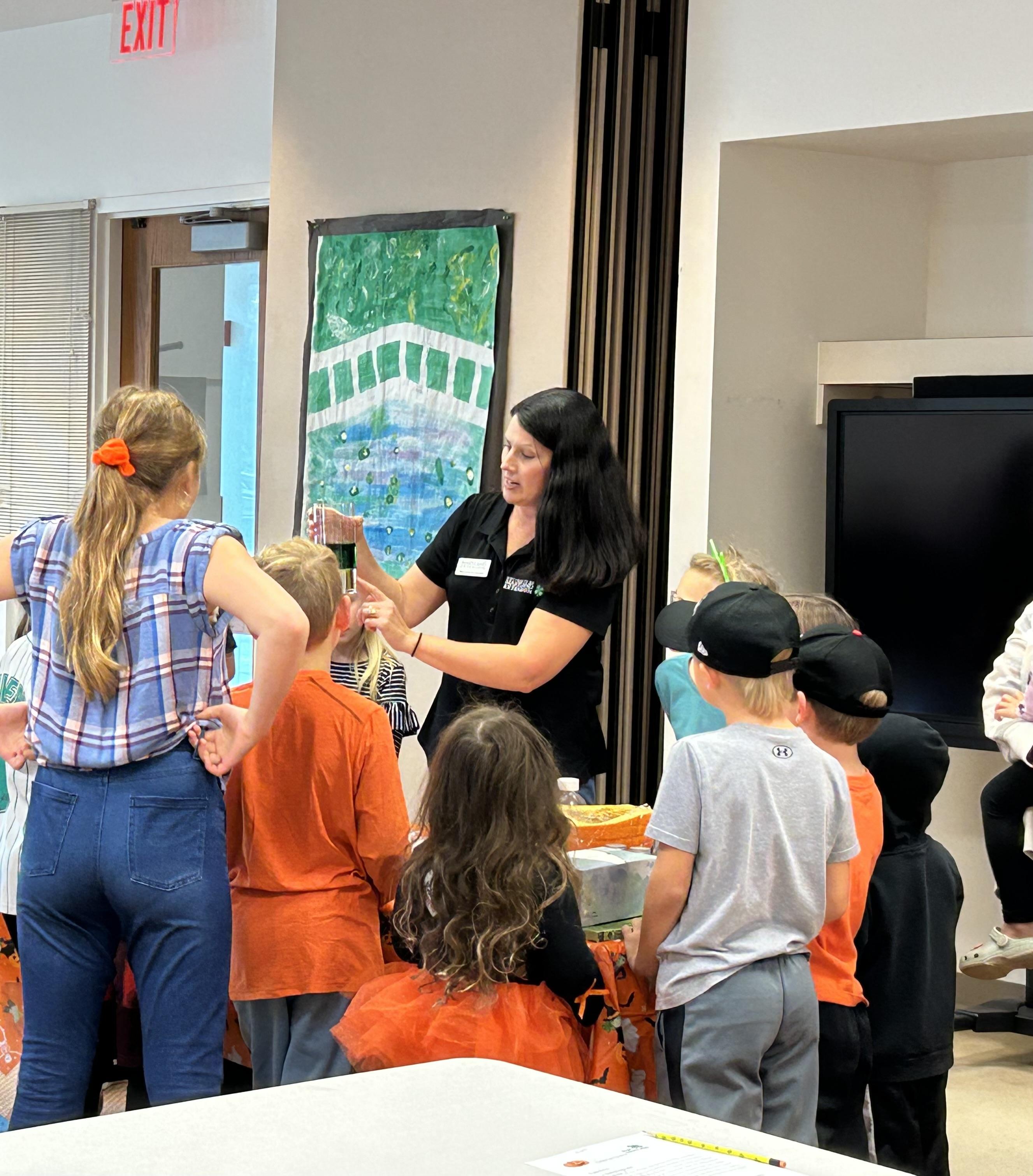 4-H educator standing at a table with students in front of her while she performs a demontration.