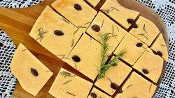 Red Lentil flatbread with rosemary and olives on top of a wooden paddle on a table