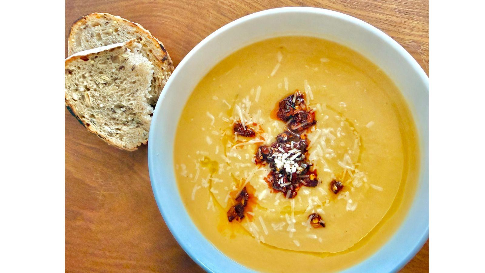 Orange soup inside a white bowl garnished with chili flakes, sitting on a wood counter with sliced bread