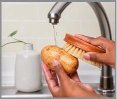 A whole potato being scrubbed under running water with a dedicated produce brush scrubbing the potato’s dimpled surface.