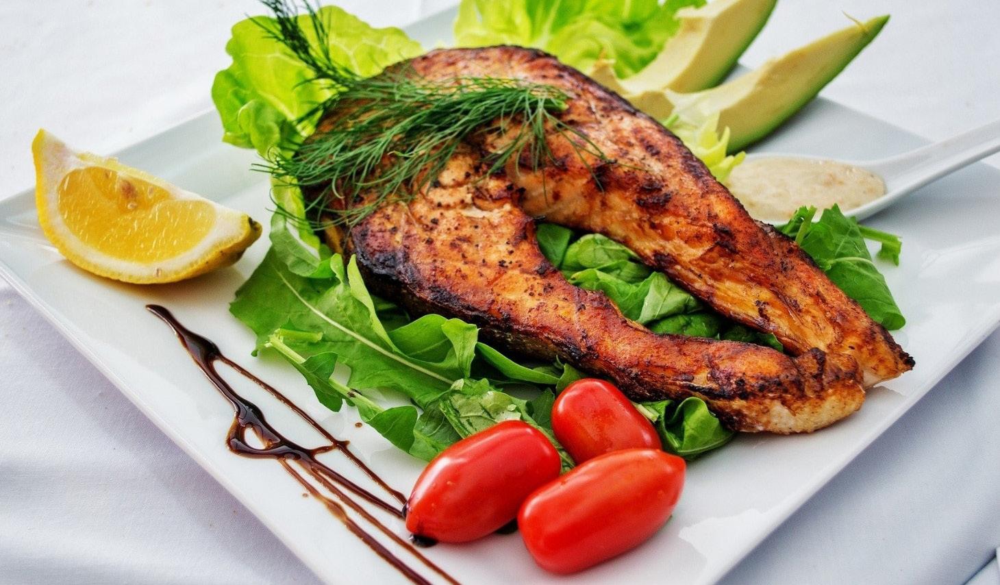 Cooked salmon accompanied by avocado wedges, three grape tomatoes, and fresh dill, all served on a bed of leafy greens and a lemon wedge.