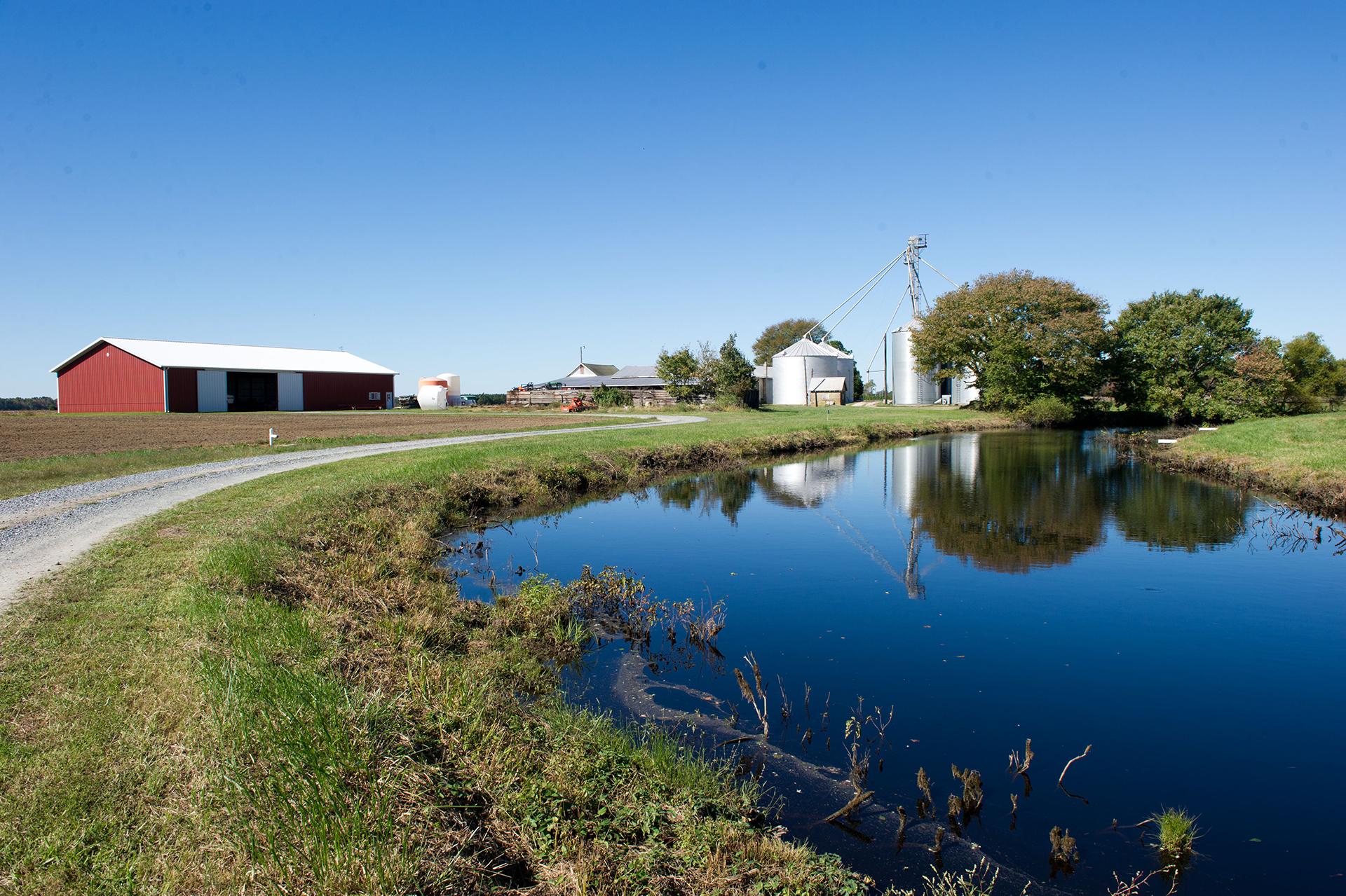 Farm land with a pond in the foreground and red barn and gray silos in the background