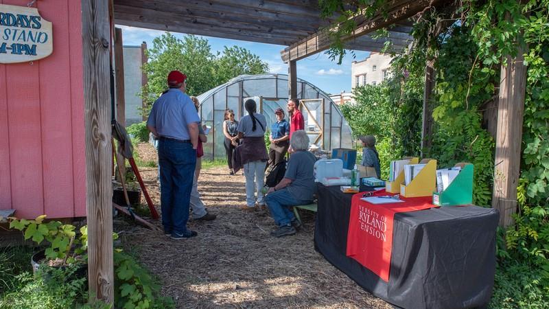 A group of people stand under a pavilion on an urban farm, near a table of educational materials