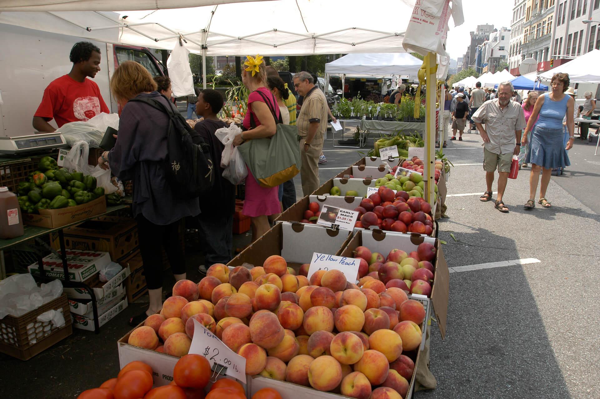 Fruit being sold at a farmers market with people paying for their produce under one of the white tents