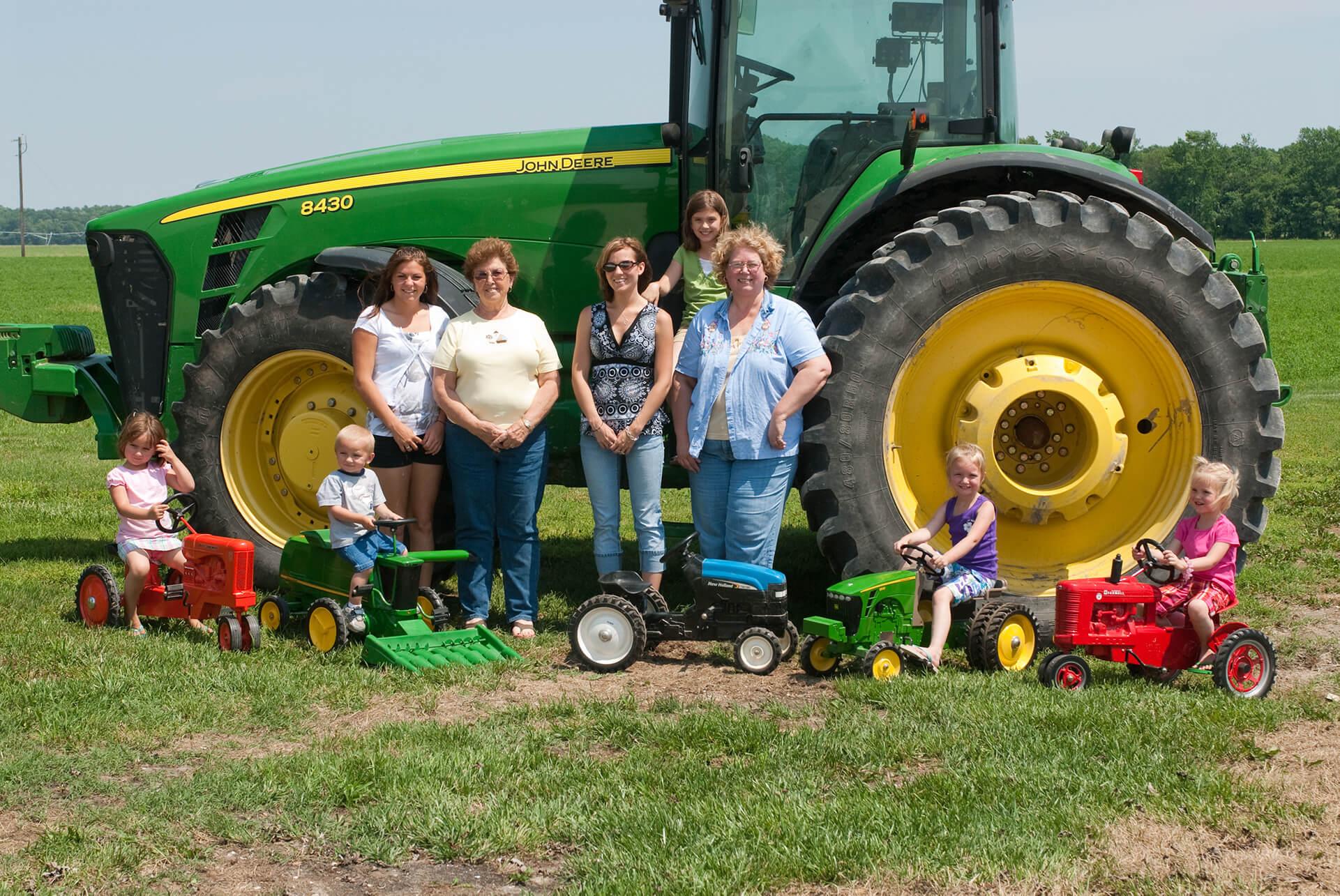 Family of women farmers standing in front of tractor with their children sitting on small toy versions of farm tractors