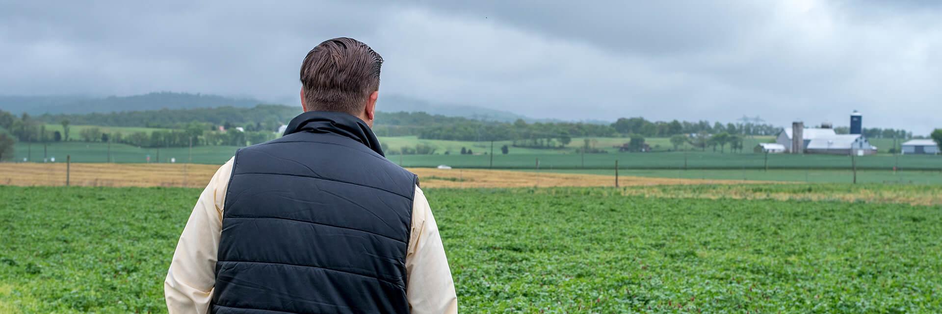 Man in black vest and yellow shirt looking off into the distance at a nearby farm with gray clouds in the background