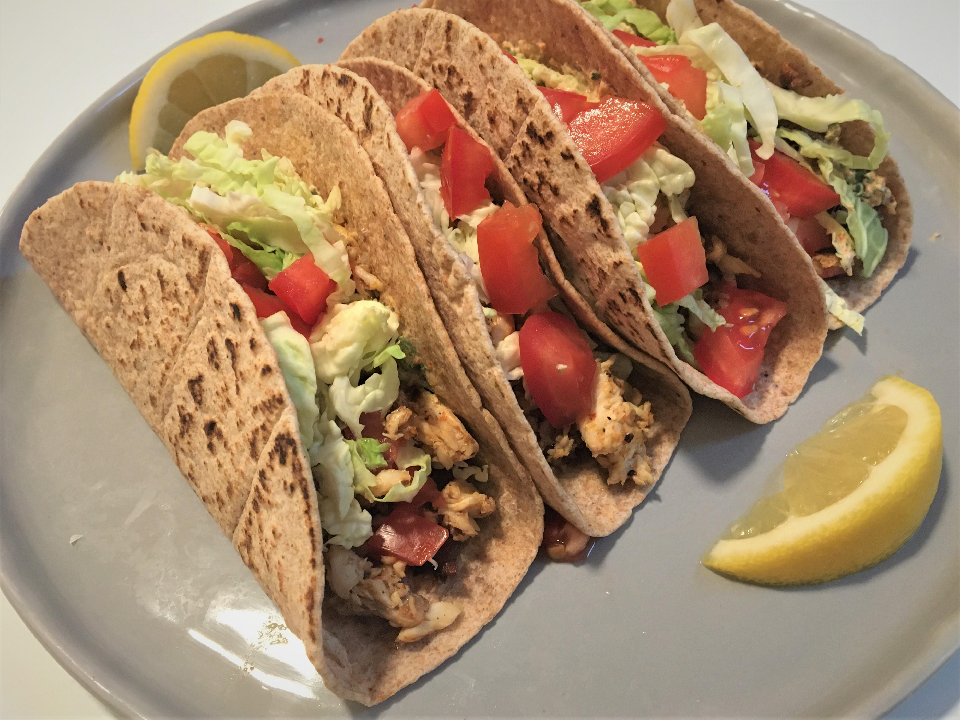 Three Whole wheat tortillas with fish, cabbage, tomatoes sitting together on a plate and garnished with lemon wedges.