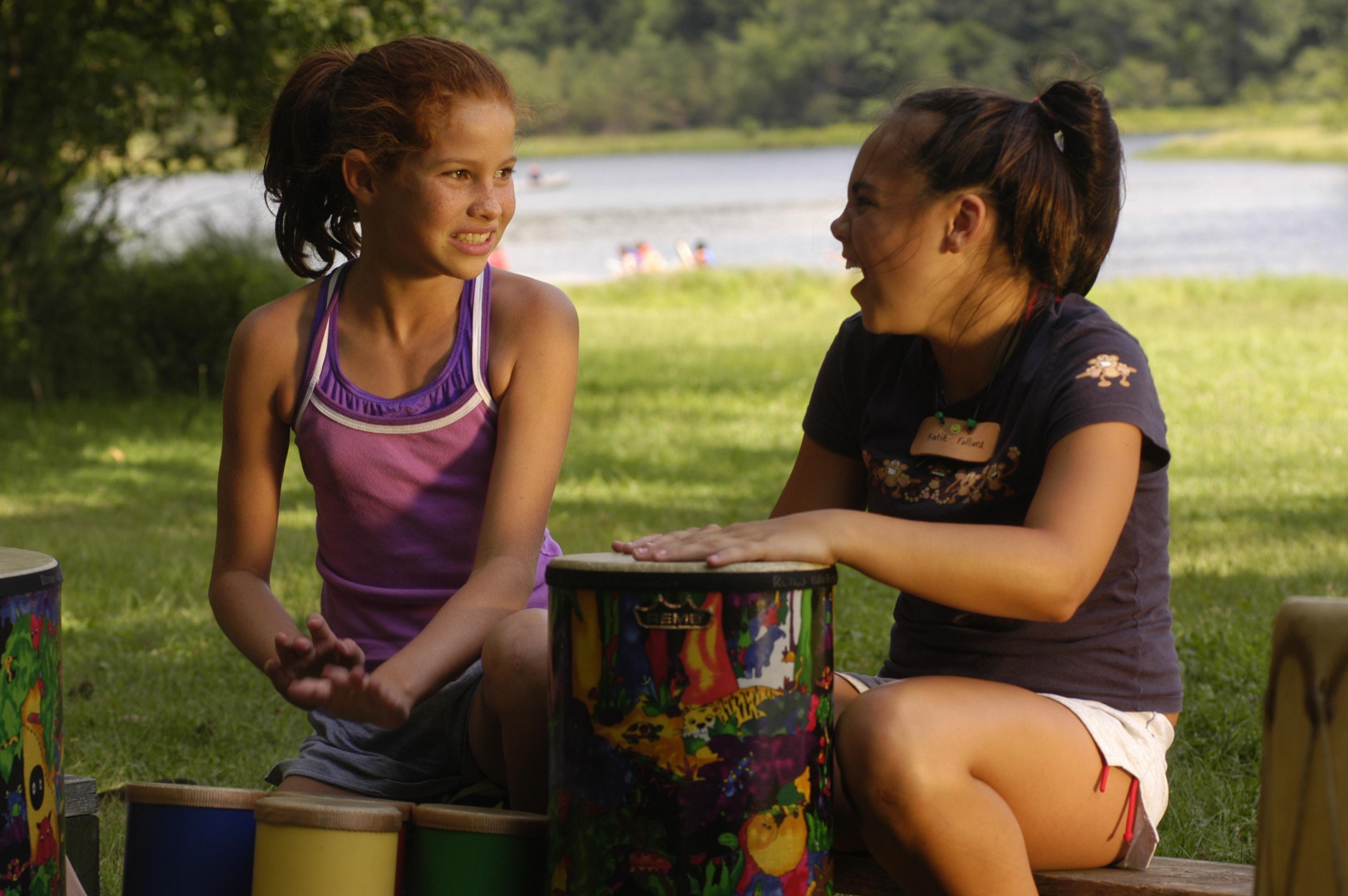 Two girls sitting on benches in front of a lake, drumming and laughing.