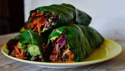 Wraps made with dark leafy greens and filled with vegetables including red cabbage and carrots stacked as 3.  Sitting on a yellow plate and a wood table.
