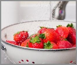 Whole strawberries being rinsed under running tap water in a colander.