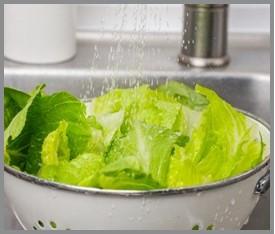 Leafy greens (lettuce) being rinsed under water in a colander.