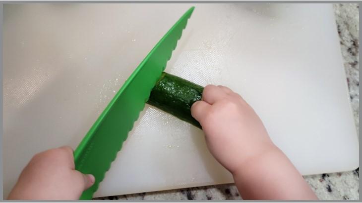 Youth chopping a cucumber that was cut lengthwise, so the cucumber has a large flat base, and cannot rock while being cut. 