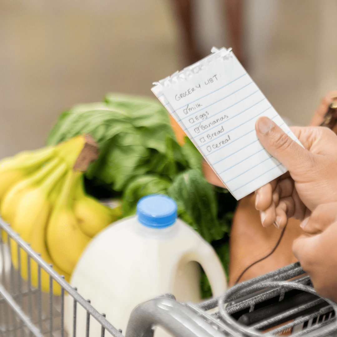 A grocery cart with bananas, spinach and gallon of milk.  An individuals hands are showing holding a grocery list that says: milk, eggs, bananas, bread, cereal.
