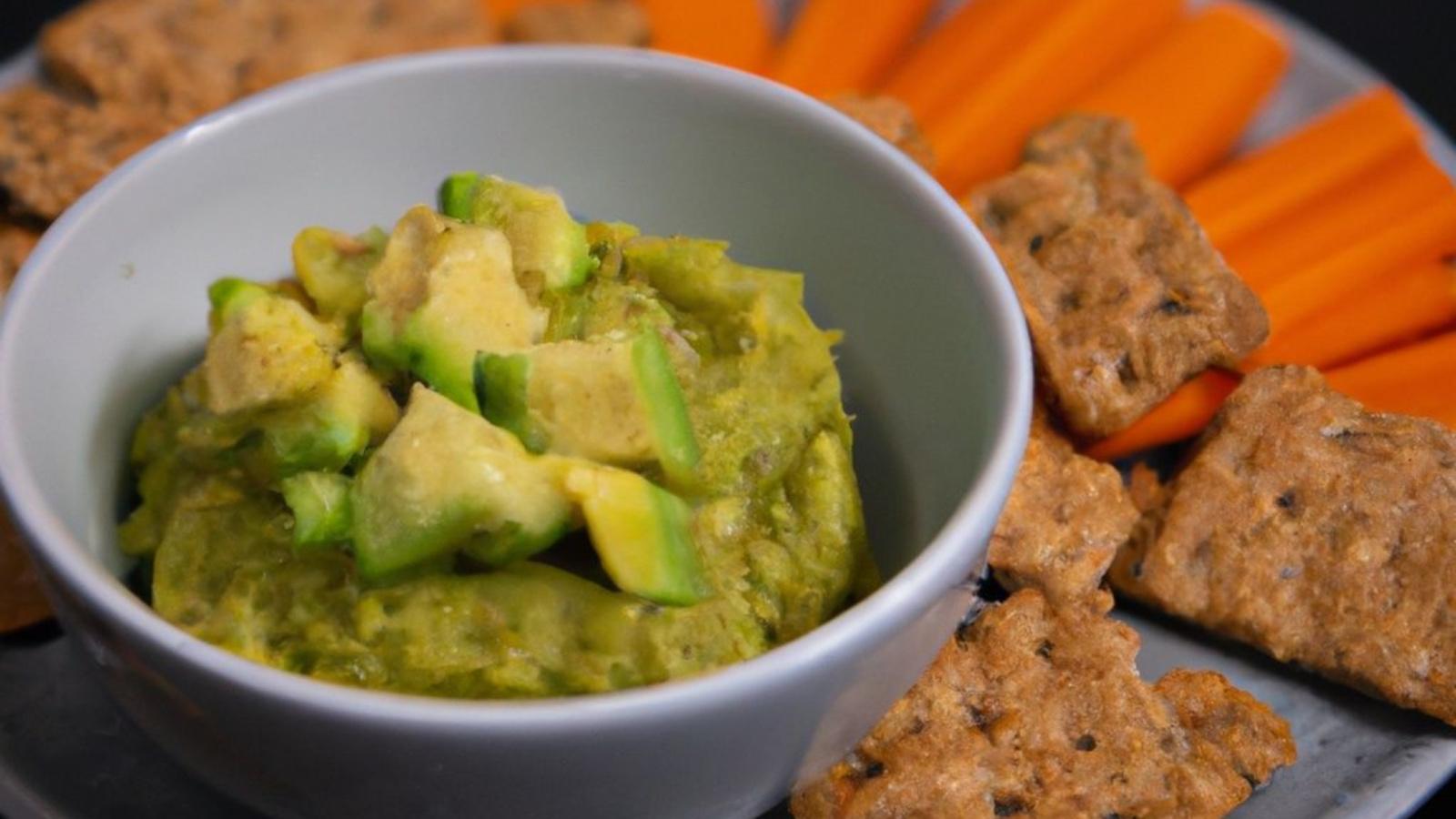A pea and avocado dip with chopped avocado on top surrounded by whole grain crackers and carrots.