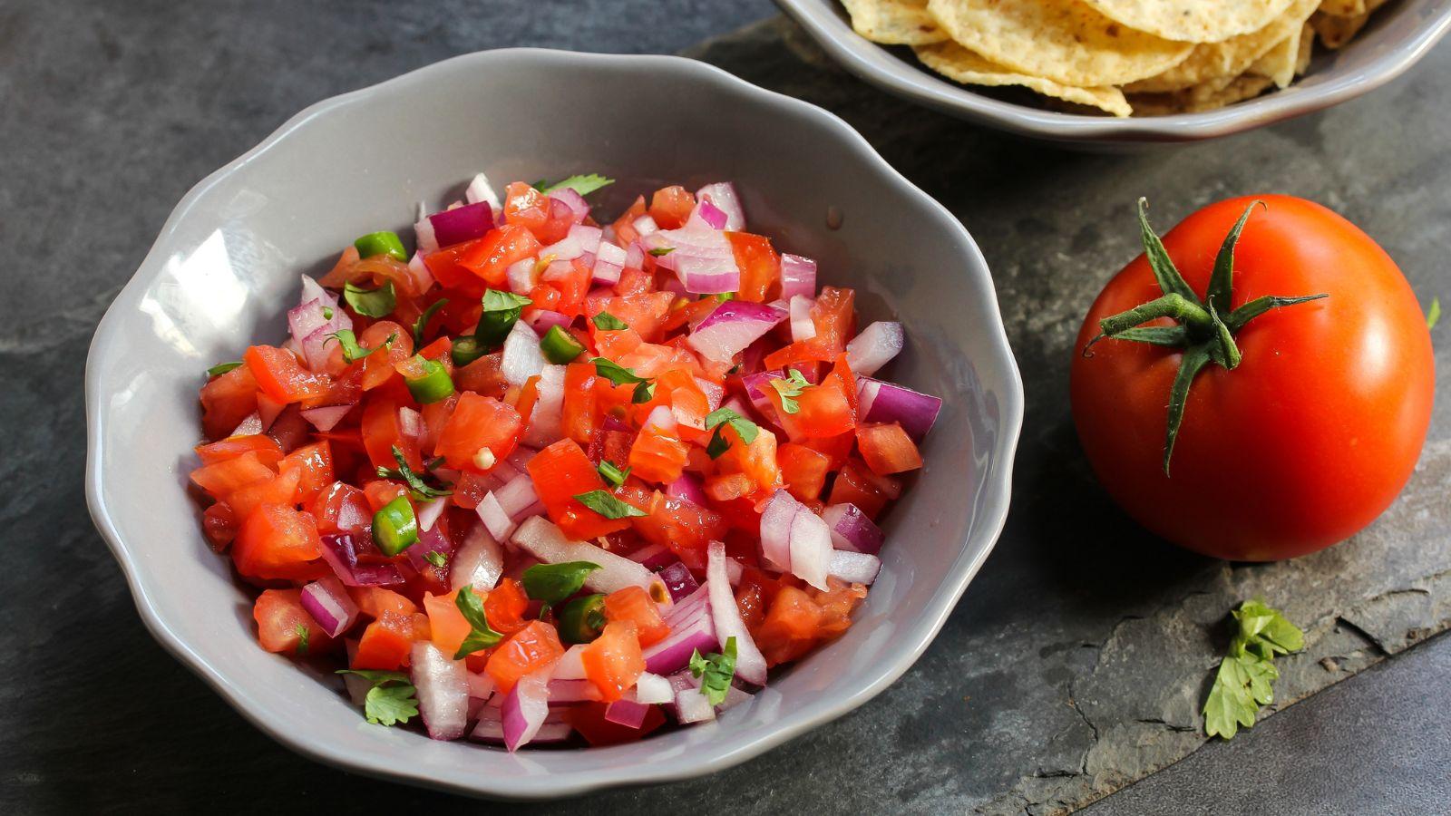 A homemade salsa wiht tomatoes, red onion, jalapeno and cilantro in a grayish bowl and a tomato to the right hand side.
