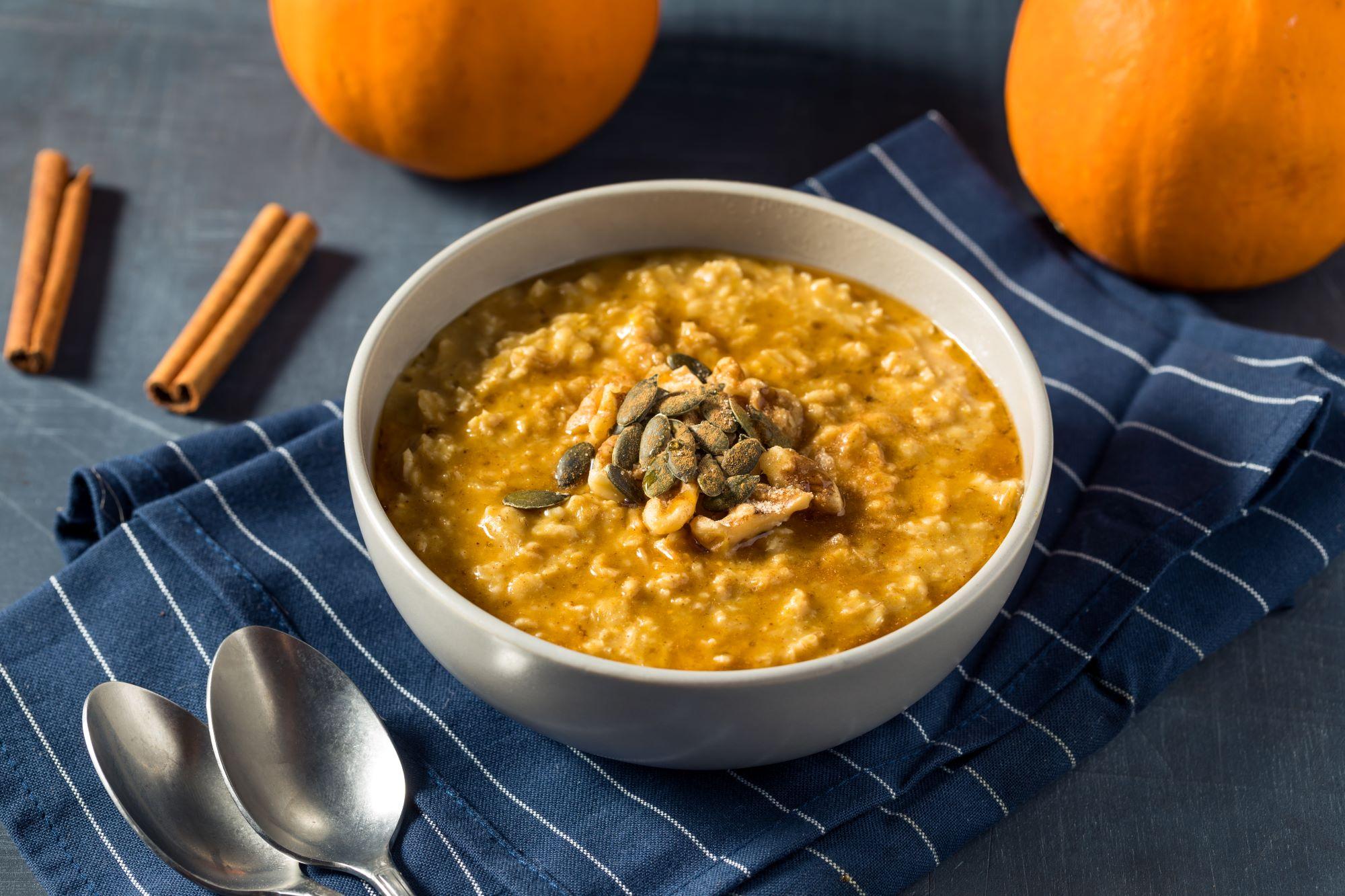 A bowl of pumpkin oatmeal topped with pumpkin seeds on a blue towel with cinnamon sticks and pumpkin in the background.