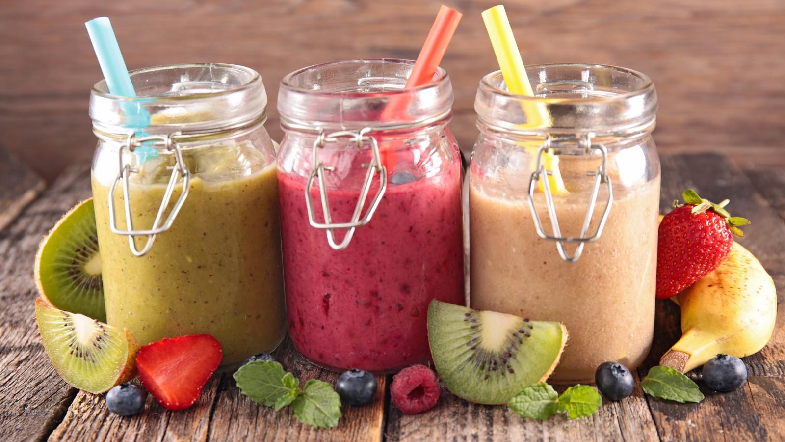 Three different kinds of smoothies - kiwi, berry, and banana with fruits in the front on a wooden background.