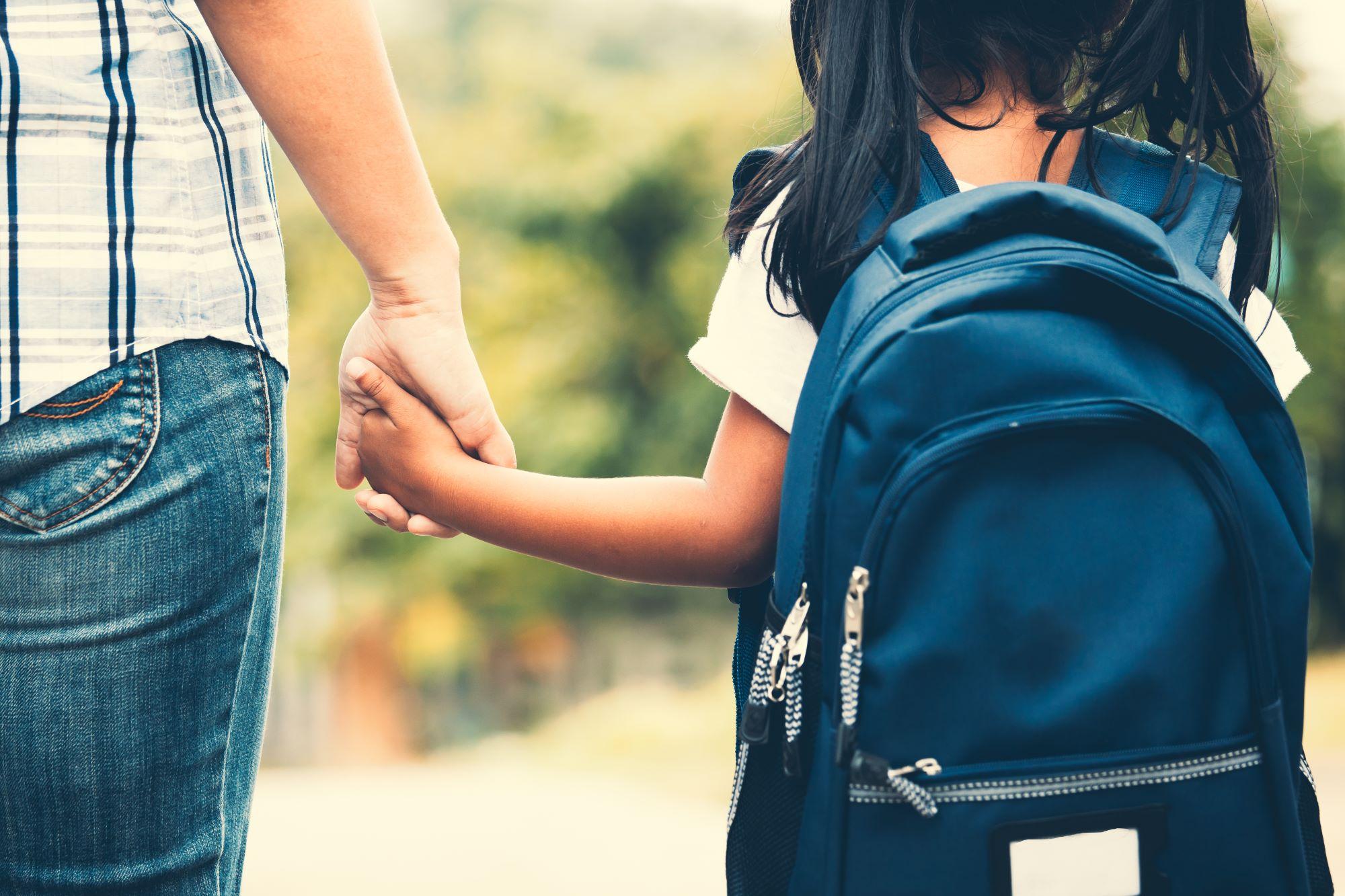 A parent and young female child with a backpack holding hands.