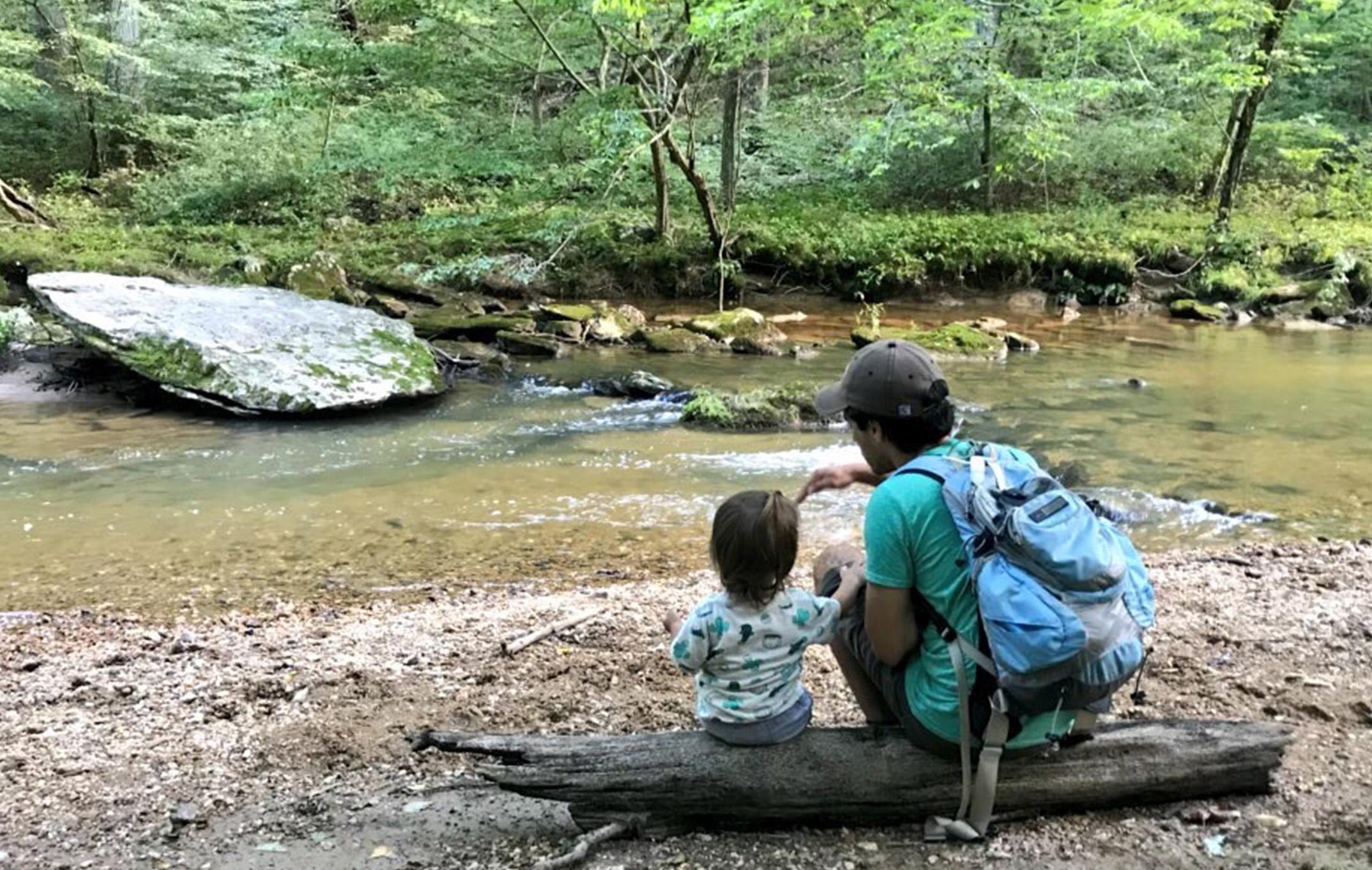  A parent and child sit on a log beside a stream and enjoy each other's company in nature's beauty.