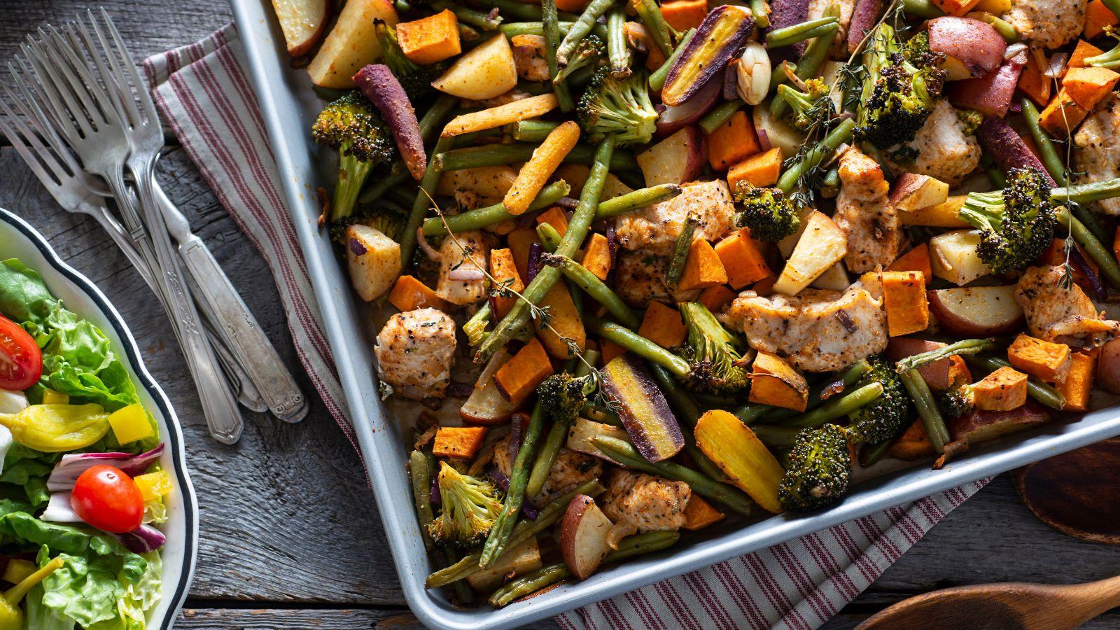 A sheet pan meal with chicken, rainbow carrots, green beans, broccoli  and carrots.  A salad on the left side with forks.