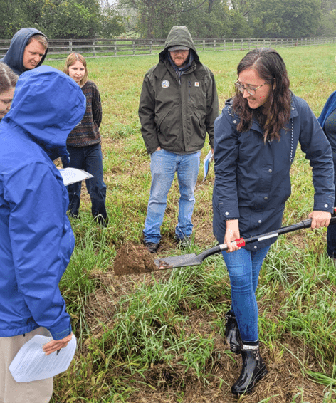 Amanda Grev, the workshop presenter, is holding a shovel of dirt in a field with the workshop participants.