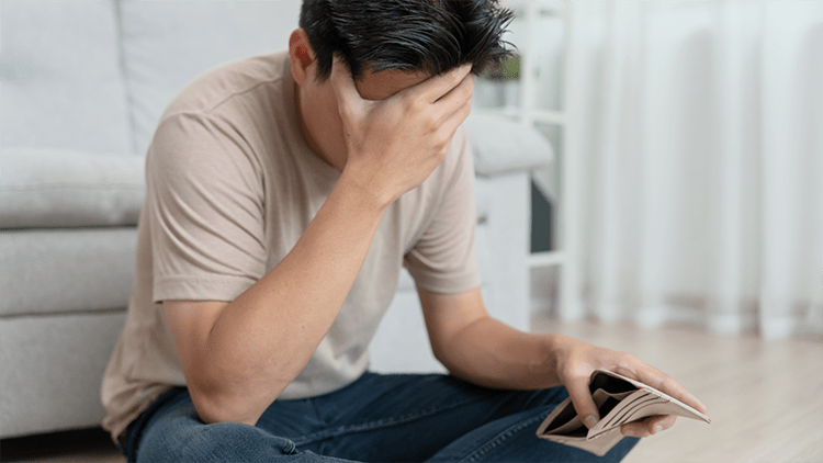 The image shows a person sitting on the floor in a living room, wearing a beige t-shirt and blue jeans. The person is holding an open wallet in one hand, which appears to be empty, while resting their head on their other hand, suggesting a sense of worry or contemplation. In the background, there is a light-colored sofa and a bright room with natural lighting.