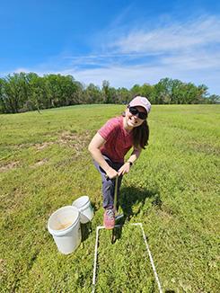 A woman in casual attire, wearing sunglasses and a cap, smiles broadly while using a tool to collect soil samples in a field. Two white buckets are nearby. Bright, sunny day.