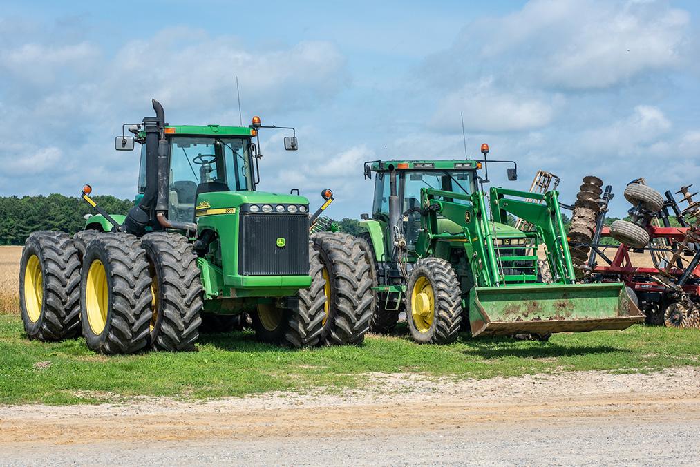 Figure 2: Two large green tractors with multiple tires are parked on grass near a dirt path under a partly cloudy sky. Farming equipment is visible in the background.