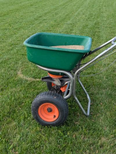 Wheeled walk-behind rotary spreader consisting of a basin that deposits grass seed on a rotating disc that flings it onto the lawn.