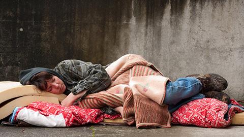  A women sleeps on a sidewalk, wrapped in blankets and a plaid shirt. They lie on red-patterned bedding, conveying a sense of vulnerability.