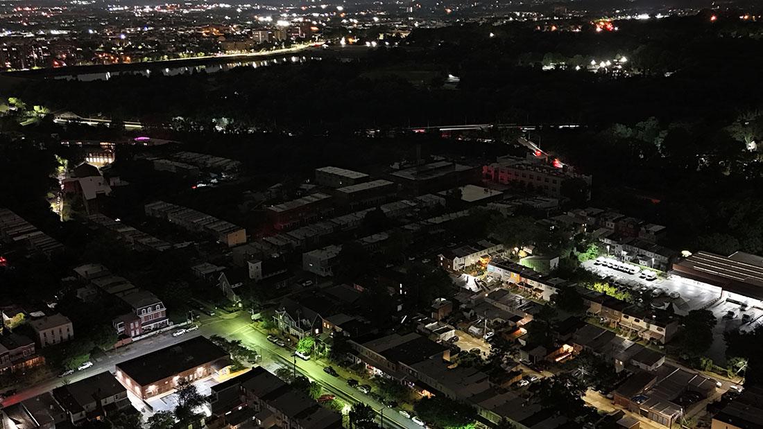 An aerial nighttime view of a city neighborhood illuminated by scattered streetlights and building lights. The lower portion shows residential blocks with rows of houses and small streets lit in greenish and white tones. To the right, a brightly lit industrial or commercial lot stands out. The center and upper areas appear much darker, with large patches of trees or undeveloped land, while the horizon shows a brightly glowing skyline filled with dense city lights.