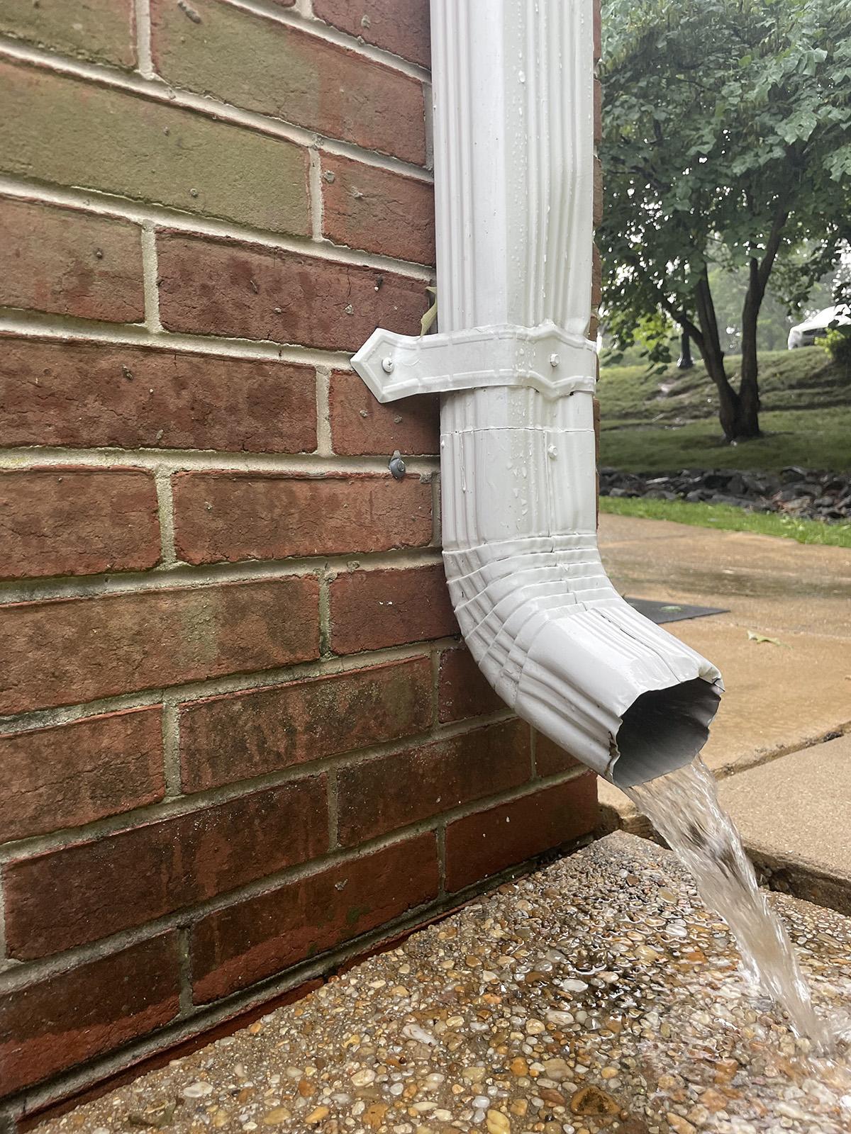 A downspout with rooftop runoff flowing out of it onto a sidewalk.