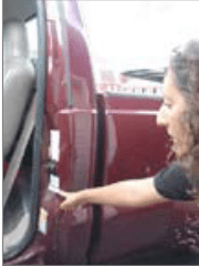 A person leaning into the open driver-side door of a maroon pickup truck points at a white and yellow information sticker on the door frame. The interior of the truck cab, including a gray seat and door latch, is visible, and the truck bed and nearby buildings appear in the background.