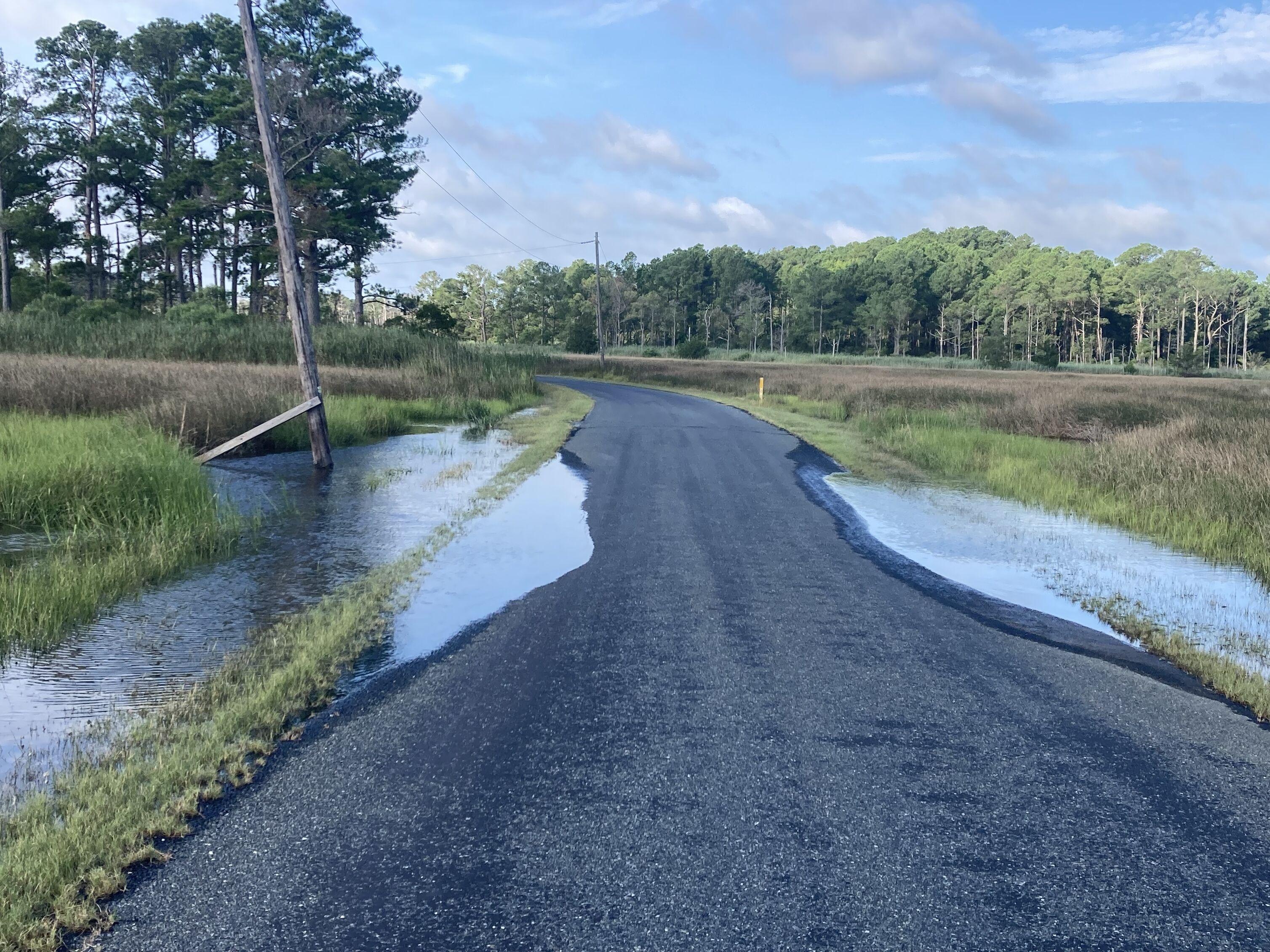A rural paved road is partially submerged by overflow from an adjacent farm drainage ditch after heavy rain, creating hazardous driving conditions.