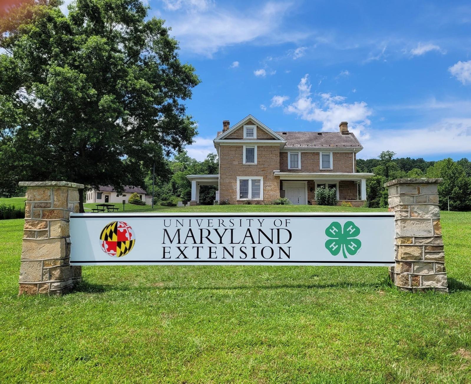 Outdoor banner sign featuring UME and 4-H logo between stone pillars with Allegany County Extension office in the background. 