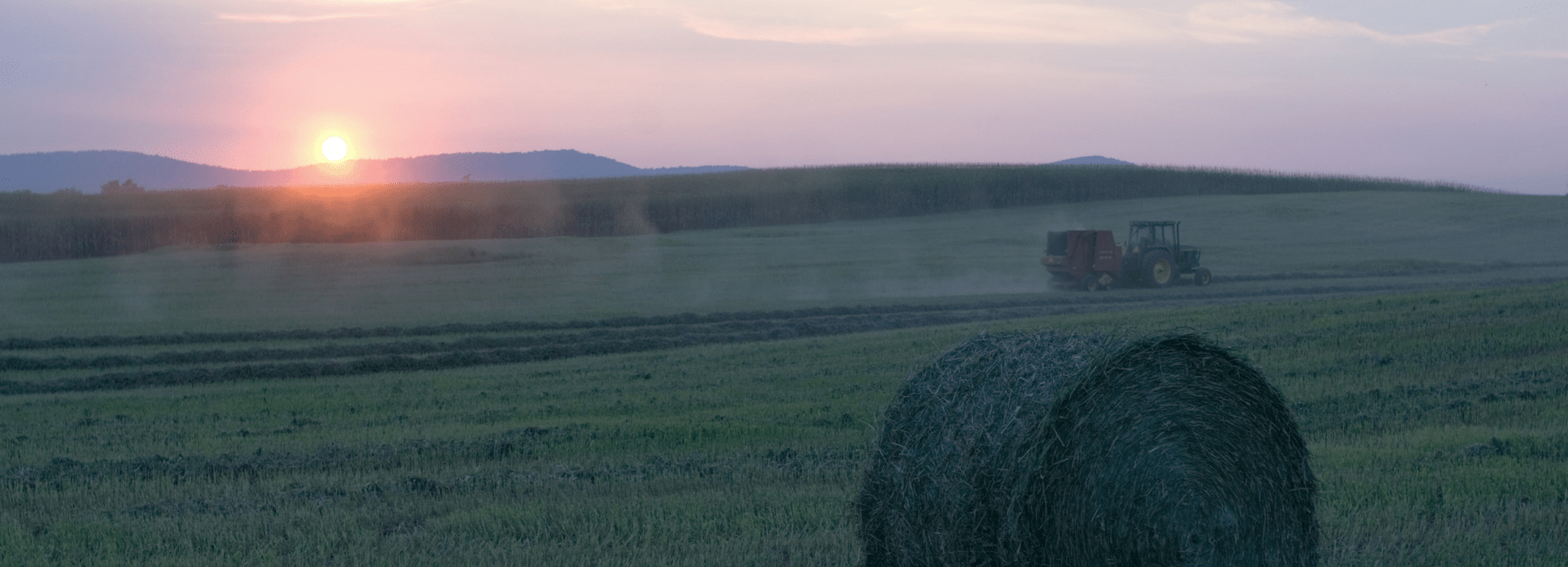 A large hay bale rests in the foreground of a cut grass field while a tractor pulls a baler in the distance under a warm sunset sky, with rolling hills and crops visible in the background.