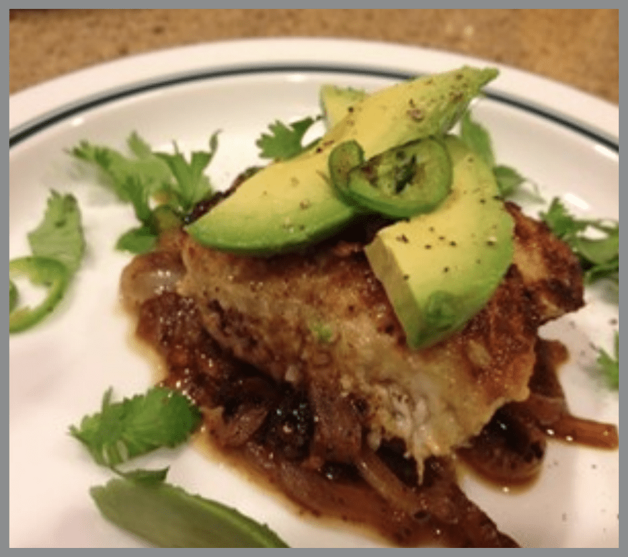 A plated dish featuring a brown, well seasoned fish fillet topped with avocado and cilantro, served on a ridged white plate.