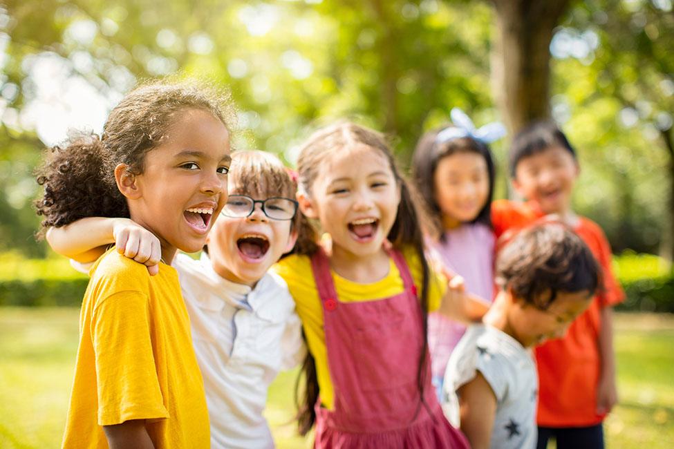 A group of six children standing close together outdoors in a sunlit park. Three children are in focus at the front, wearing bright yellow, white, and pink clothing, with arms around one another's shoulders. Three more children stand slightly behind and to the side, blurred due to shallow depth of field.