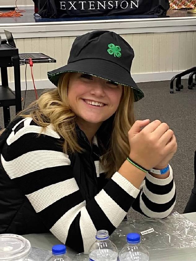 Person wearing a black bucket hat with a green 4-H clover logo sits at a table indoors, hands clasped together. The person is wearing a black-and-white striped sweater and has shoulder-length hair visible beneath the hat.