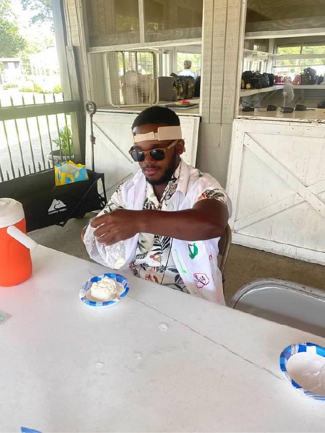 Person wearing sunglasses, a patterned shirt, and a white vest sits at a table indoors while handling a plastic bag near a paper plate with whipped cream or foam. A beige strap is wrapped around the person’s head. An orange water cooler sits on the table, and shelves, windows, and bags are visible in the background of the open-air building.