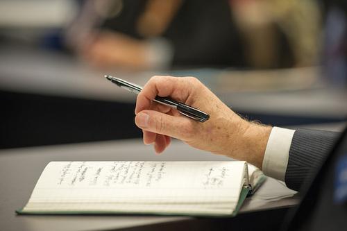 Close-up of a person’s hand holding a pen above an open notebook with handwritten notes. The notebook rests on a table in a meeting or conference setting, with a blurred background of attendees and seating.