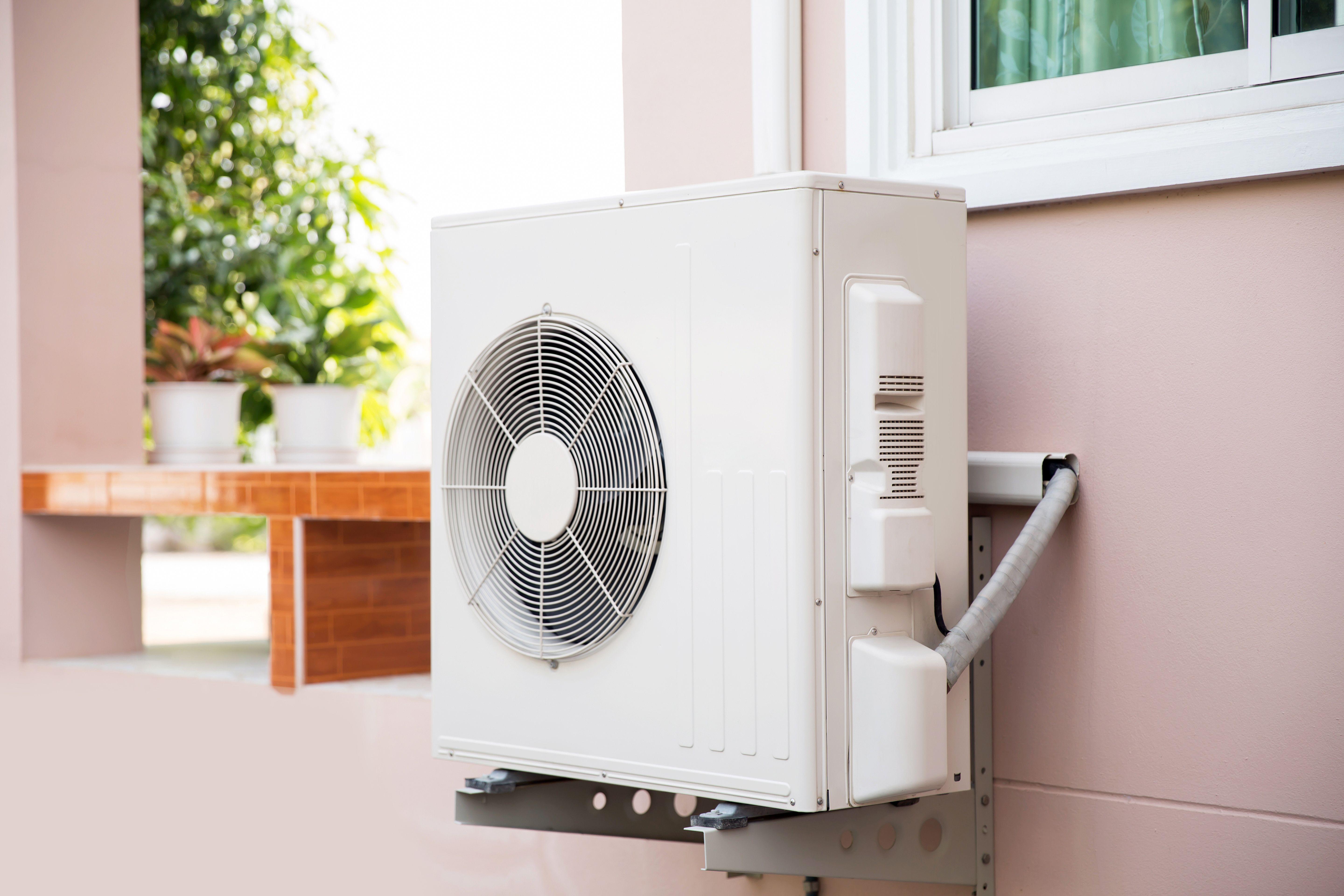 outdoor air‑source heat pump unit mounted on a residential wall, showing fan grille and refrigerant lines connected to the home