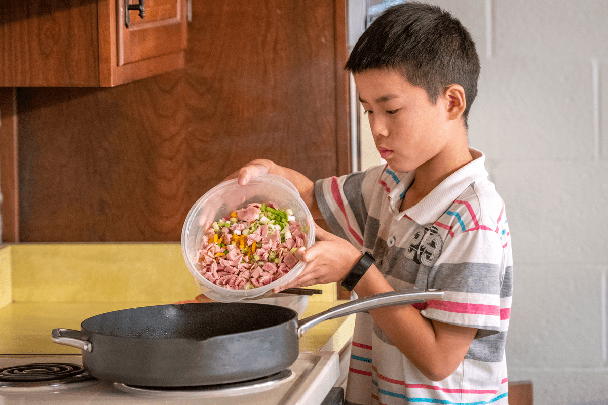 a young boy pouring a mixture of chopped vegetables and meat from a plastic container into a frying pan on a stovetop.