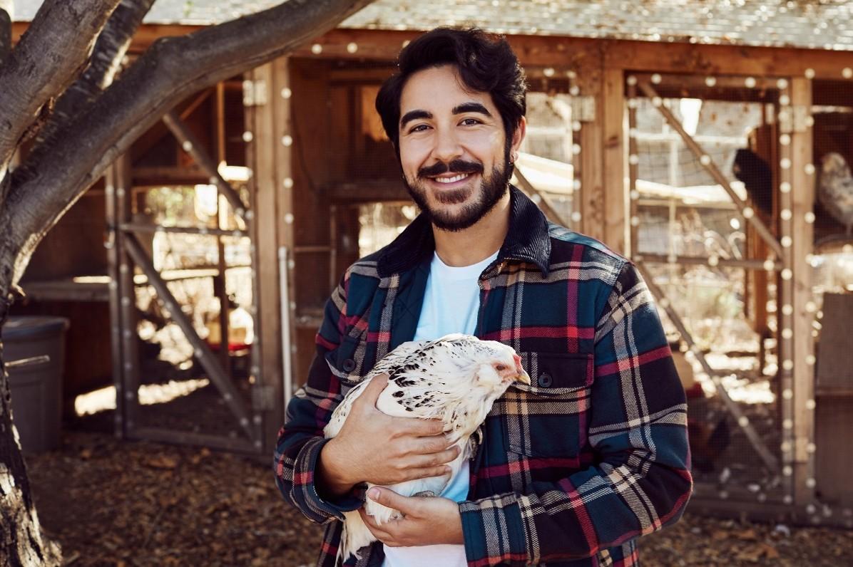 A person standing outdoors in front of a wooden chicken coop holds a white chicken with black speckles. The person is wearing a plaid flannel-style jacket over a light-colored shirt. Trees cast shade across the scene, and additional chickens are visible inside the coop in the background. Warm natural light illuminates the yard and wooden structures.