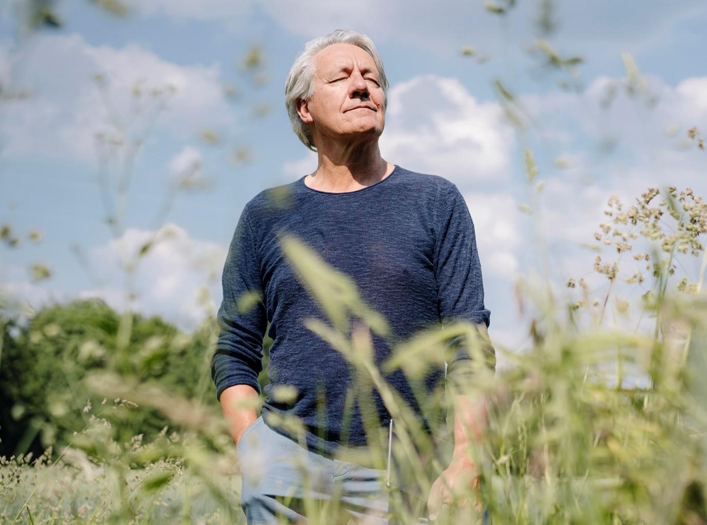 A person with gray hair stands in a grassy meadow under a partly cloudy sky. The person is wearing a long-sleeved dark blue shirt and light-colored pants. Tall grasses and wildflowers are visible in the foreground and background, with the person positioned near the center of the image. The photo is taken from a low angle, and the surrounding vegetation is softly blurred.