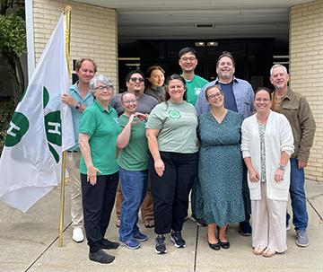 Group photo of approximately 11 people standing outside a brick building entrance next to a white 4-H flag. Several participants are wearing green 4-H shirts, including one shirt displaying the "Beyond Ready" logo. The group is posed on a sidewalk in front of the building.