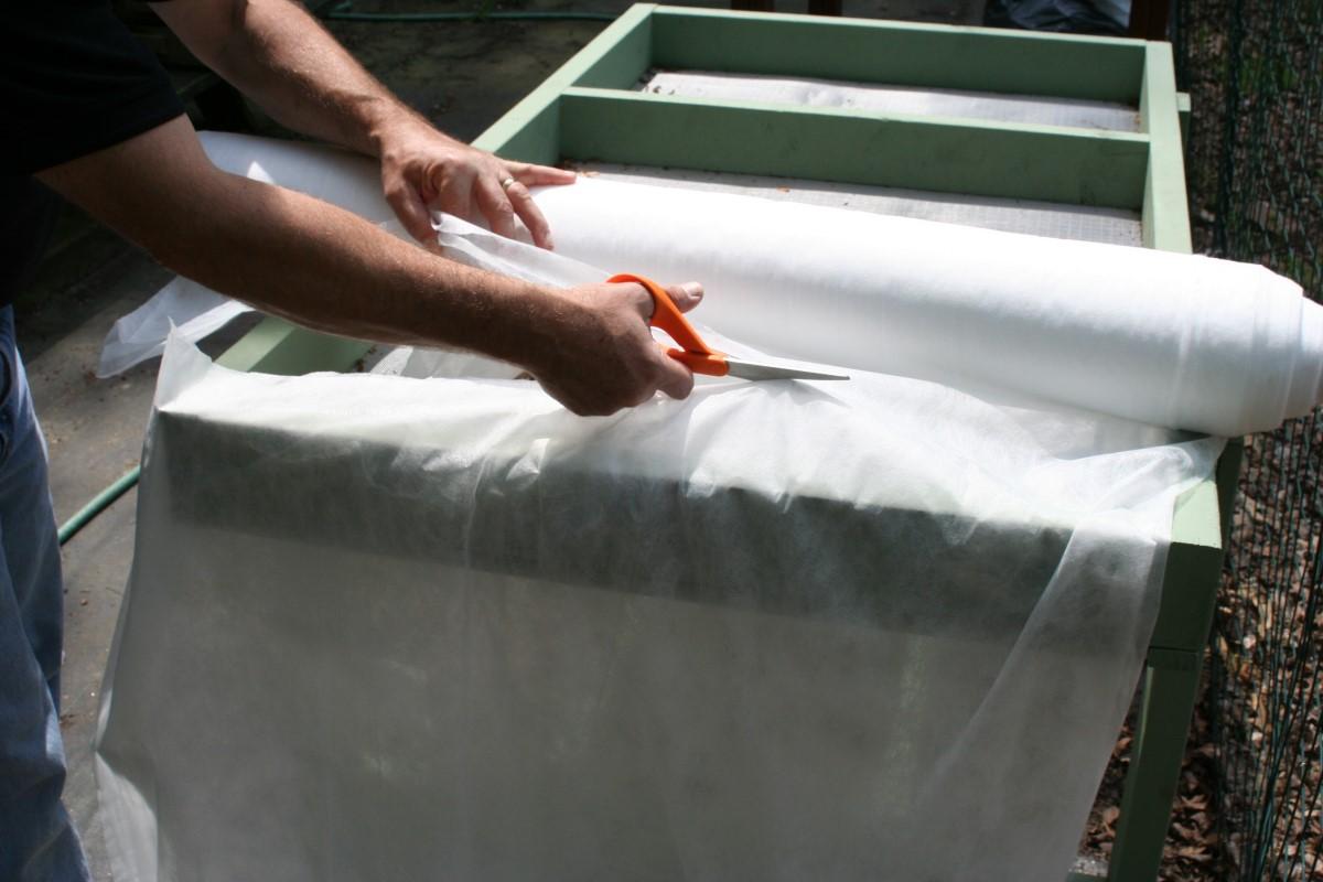 A person uses scissors to cut a length of row cover material from a fabric roll 7 feet wide set on top of an empty salad table.
